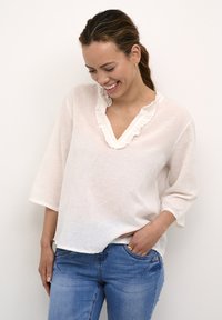 Bluză albă din in cu decolteu volan și mâneci trei sferturi, purtată cu blugi albaștri. Bluza are o croială lejeră și o textură subtilă.