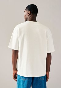 Kurzärmeliges, cremefarbenes Hemd mit rundem Ausschnitt, lockerer Passform und weicher Textur, kombiniert mit blauen Jeanshosen.