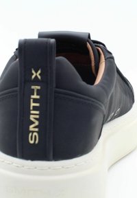 Sneaker in pelle nera con suola in gomma bianca, dotata di colletto imbottito e logo dorato sul tallone. Design minimalista con linee eleganti.