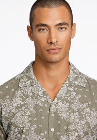 Camisa verde de manga corta con patrones de paisley blancos, cuello de punta y cierre de botones. Fabricada con un tejido ligero y un acabado texturizado.