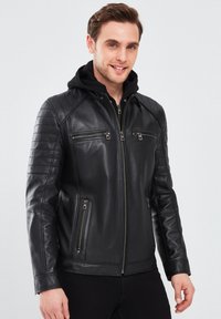 Chaqueta de cuero negra con un diseño ajustado, cuello con capucha y acentos acolchados en los hombros. Cuenta con bolsillos frontales con cremallera y una textura suave.