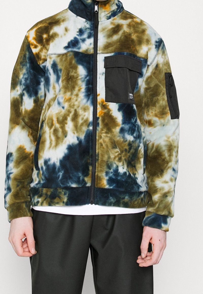 Personne portant une veste polaire zippée avec un motif tie-dye bleu, vert et marron et des poches zippées noires sur la poitrine et la manche.