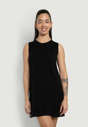 Donna con lunghi capelli scuri in un abito nero senza maniche, sorridente, con tatuaggio floreale sull'avambraccio destro su sfondo semplice.