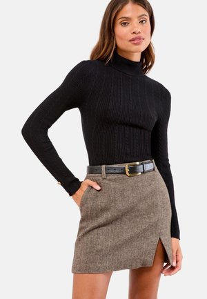 Femme portant un pull à col roulé noir en maille torsadée et une mini-jupe en chevrons marron avec une ceinture noire, posant avec une main dans la poche.