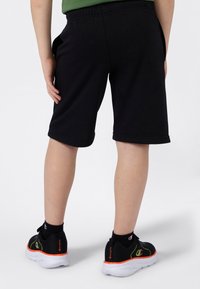 Enfant portant un short noir jusqu'aux genoux, des baskets noires avec des détails orange et vert, et des chaussettes noires, se tenant sur un fond blanc.
