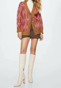 Cardigan marron avec un motif losange rose et orange, boutonné devant, associé à une jupe marron et des bottes beige mi-mollet à talons blocs.