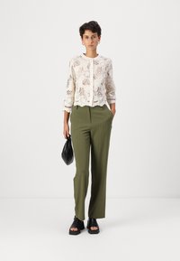 Blonde, langermet, hvit bluse med blomstermønster og rund hals, kombinert med olivengrønne stripete bukser og svarte platåsandaler.