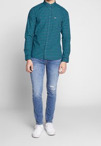 Camicia a maniche lunghe a quadretti verde acqua e nera con chiusura a bottoni, abbinata a jeans strappati azzurri chiari e sneaker bianche.