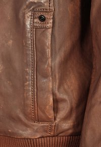 Sectie van een bruine leren jas met een zijzak en een zwarte drukknoop. Het materiaal heeft een gestructureerde, licht verweerde afwerking.