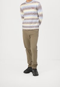 Pull rayé avec des nuances de bleu clair, marron et jaune ; texture côtelée. Associé à un pantalon beige et des chaussures de sport noires.