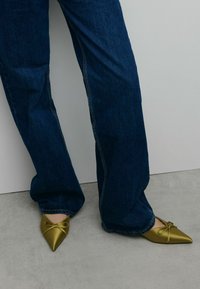 Ballerines en satin vert olive à bout pointu avec un détail en nœud. Portées avec un jean bleu foncé évasé, sur un fond neutre.