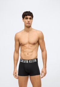 Barbröstad man med kort lockigt hår iförd svarta Calvin Klein boxershorts, stående framför en ljusgrå enkel bakgrund.