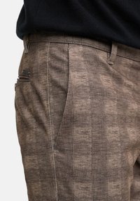 Pantalones marrones de estampado de cuadros con bolsillos laterales, usados con una camiseta negra, mostrando la cintura y la parte superior del muslo.