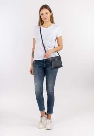 Black crossbody bag with zipper, worn over a white t-shirt and distressed blue jeans. Model has straight hair and white sneakers.