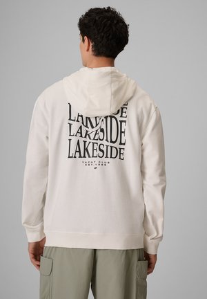 Mężczyzna w białej bluzie z kapturem z czarnym napisem „Lakeside Yacht Club Est. 1995” z tyłu i jasnych zielonych spodniach typu cargo, zwrócony tyłem.