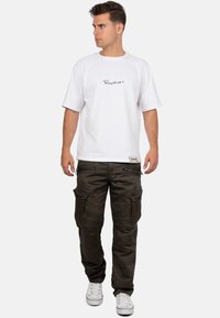 Junger Mann in einem weißen kurzärmeligen Hemd und dunklen Cargo-Hosen mit weißen Turnschuhen, der steht und zur Seite schaut.