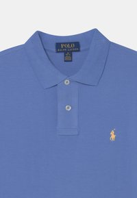 Ljusblå polo skjorta med två knappar och en liten gul logotyp av en ryttare på vänster bröst. Tydlig textur och detaljer på kragen synliga.