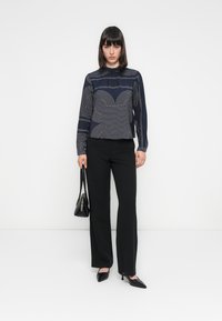 Marinebluse mit weißen geometrischen Linienmustern, hochgeschlossen, mit langen Ärmeln. Schwarze, maßgeschneiderte Hosen, spitze Absätze und eine kleine schwarze Tasche.