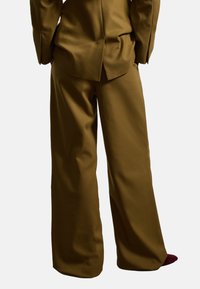 Pantaloni larghi marroni realizzati in tessuto morbido, caratterizzati da una vestibilità sartoriale e pieghe sottili. Il cinturino è piatto e non ci sono tasche visibili.