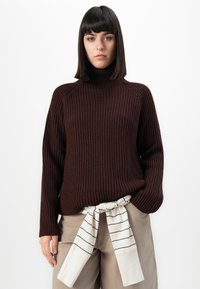 Frau mit glattem schwarzen Haar, die einen dunkelbraunen gerippten Rollkragenpullover und beige Hosen trägt, mit einem gestreiften Hemd, das um die Taille gebunden ist.