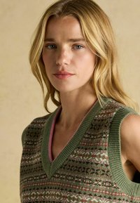 Blond naine kerge meigiga, seljas roheline, roosa ja pruuni mustriga kudumita ilma varrukateta sviiter, taust ilma mustrita.