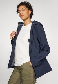 Marineblå vandtæt jakke med hætte, lynlås foran og elastiske manchetter, båret over en hvid t-shirt og olivengrønne cargo bukser.