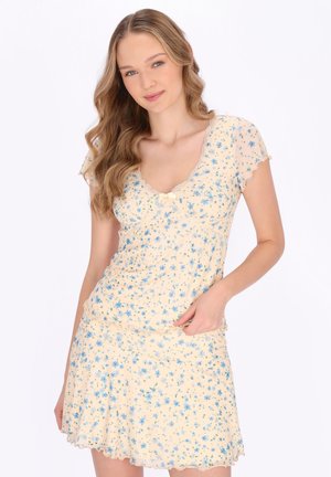 Jonge vrouw met golvend blond haar, gekleed in een crème jurk met blauwe bloemenprint en kanten afwerking, poseert met één hand op haar heup.