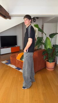 Mladý muž v čiernom tričku a nadrozmerných sivých džínsoch stojí v modernom obývacom priestore s oranžovou pohovkou, televízorom a veľkou zelenou rastlinou.