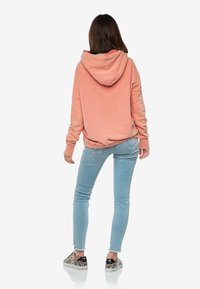 Felpa con cappuccio color pesche, dal tessuto morbido, vestibilità ampia e polsini a costine, abbinata a jeans skinny azzurri e sneaker con motivo.