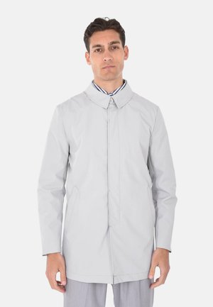 Uomo che indossa un cappotto lungo grigio chiaro con bottoni e una camicia con colletto a righe, su sfondo bianco, rivolto verso avanti con espressione neutra.