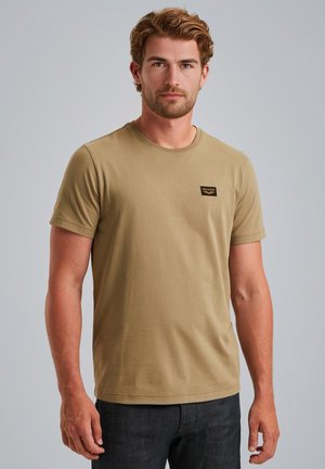 T-shirt kaki à manches courtes en coton doux et extensible, avec un col rond et une petite étiquette noire cousue sur la poitrine. Porté avec un jean foncé.