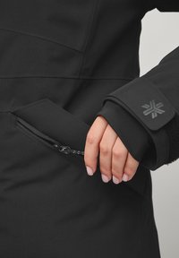 Zwarte jas met een gladde textuur, voorzien van een ritszak en een manchet met textuurelementen. Het ontwerp is minimalistisch en functioneel.