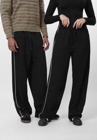 Két személy fekete, széles szárú nadrágot visel, fehér oldalsávval és fekete sportcipőben, egyikük a nadrág zsinórját igazítja.