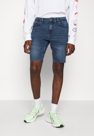 Pantalones cortos de mezclilla en azul oscuro, con un acabamiento deshilachado y diseño clásico de cinco bolsillos. Combinados con una camiseta de manga larga blanca y zapatillas verdes.