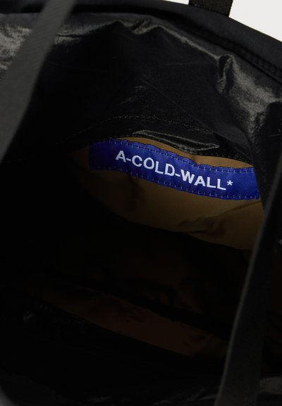 Unutrašnjost crne platnene torbe s plavom etiketom s belim tekstom koji glasi "A-COLD-WALL*" i glatkim smeškim podstavom.