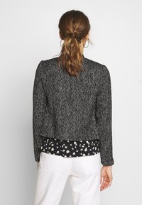 Sort-hvid sildebensjakke med strukturerede skuldre. Tekstureret stof, kort design, båret over en blomsterbluse. Hvide bukser synlige.