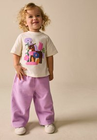 Světlounce žluté tričko s barevným motivem poníka a květin, doplněné volnými levandulovými kalhotami a bílými botami. Celkově hladká textura.