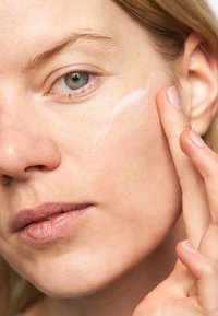 Närbild av en person som applicerar en genomskinlig kräm på sin kind. Huden ser slät ut med naturliga toner, vilket framhäver en hudvårdsrutin.