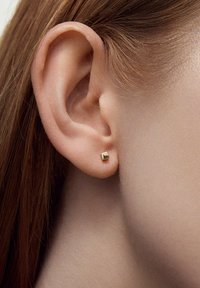 Złoty kolczyk w kształcie geometrycznego kwadratu, z polerowaną powierzchnią. Noszony na małżowinie usznej, dopełnia naturalne włosy.