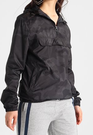 Sort camouflagemønstret vindjakke med halv lynlås, frontlomme med klap, elastiske manchetter, båret med lysegrå bukser med marineblå sidelommer.