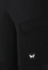 Svarta skidbyxor med texturerad yta, som har en stor cargoficka, förstärkta sömmar och en logotyp som säger "WHISTLER."