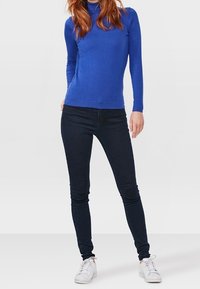 Un pull-ras du cou à manches longues bleu associé à un jean skinny noir et des baskets blanches. Le pull présente une texture lisse et un design ajusté.