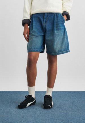Person trägt blaue Denim-Shorts, weiße Socken, schwarze Puma-Sneaker und einen cremefarbenen Pullover mit grauen Bündchen und steht auf einem blauen Teppich.