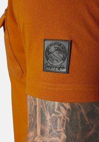 Orange cotton shirt with a textured finish. Features a black rectangular patch displaying mountain graphics and brand name.