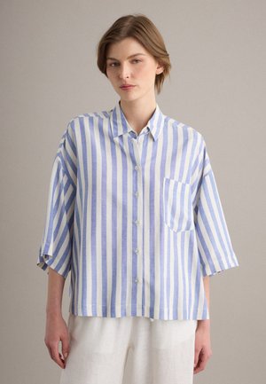 Persona joven con camisa suelta de rayas verticales azules y blancas con bolsillo delantero y pantalones blancos, de pie frente a un fondo liso.