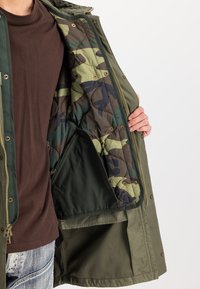 Άτομο που φοράει ελαιοπράσινο μπουφάν ανοιχτό ώστε να φαίνεται η επενδυτική καπιτονέ επένδυση σε καμουφλάζ και η εσωτερική τσέπη, ντυμένο με καφέ πουκάμισο και μπλε τζιν.