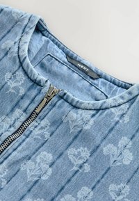 Vaatehihna vaaleansinisessä denimissä, jossa on sävyton kukkakuosi. Varustettu vetoketjulla ja palttinakuosisella pinnalla sekä pyöreällä kaula-aukolla. Brändilogo näkyvissä.