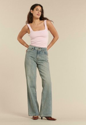 Kobieta o długich ciemnych włosach, ubrana w jasnoróżowy bezrękawnik, jeansy z wysokim stanem i szerokimi nogawkami oraz brązowe szpiczaste szpilki, stoi z rękami na biodrach.