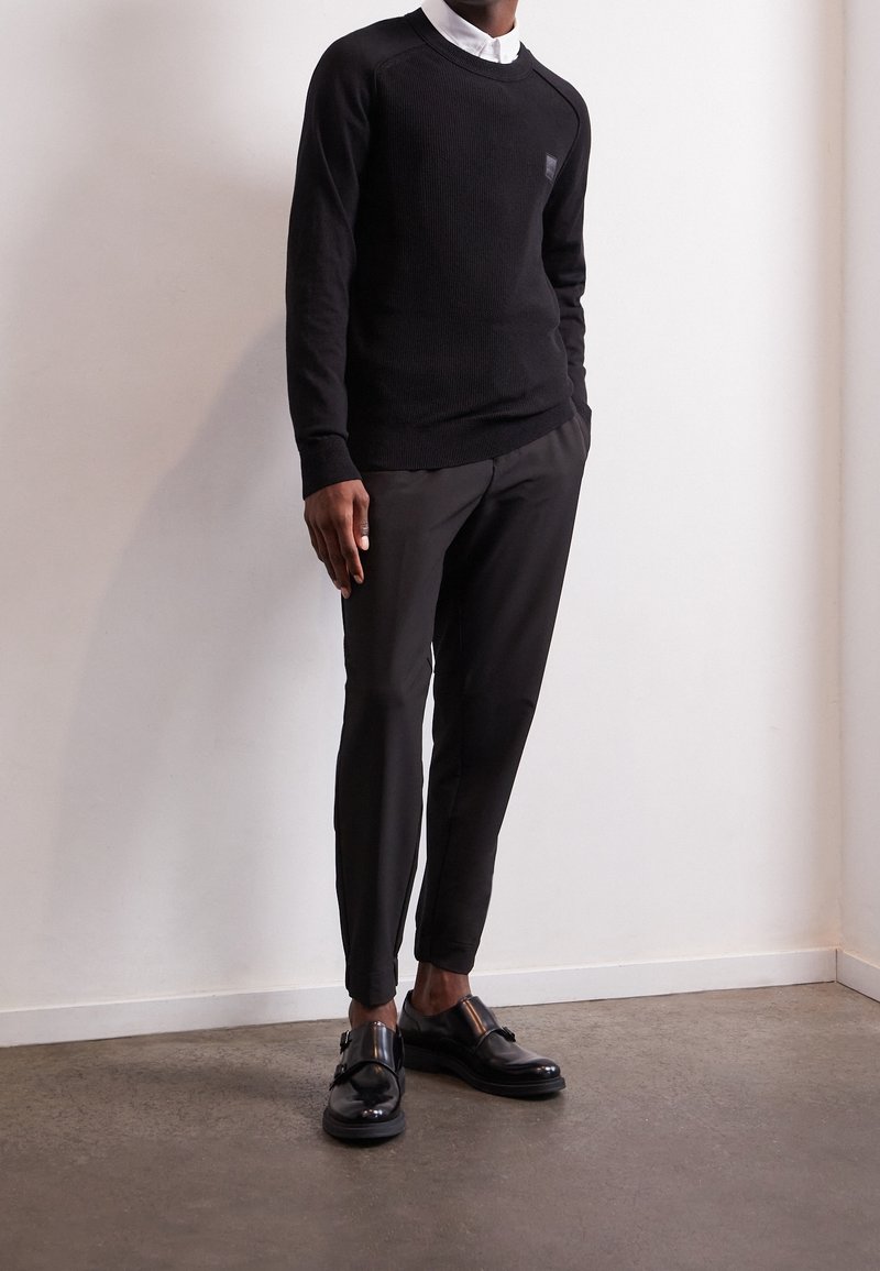 Svart stickad tröja med rund halsringning, kombinerad med skräddarsydda svarta byxor och blanka svarta skor. Släta texturer och figursydd design.