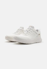 Baskets de sport blanches avec un dessus en tissu texturé, fermeture à lacets et semelle rembourrée ; se caractérisent par un accent de logo sur le côté.